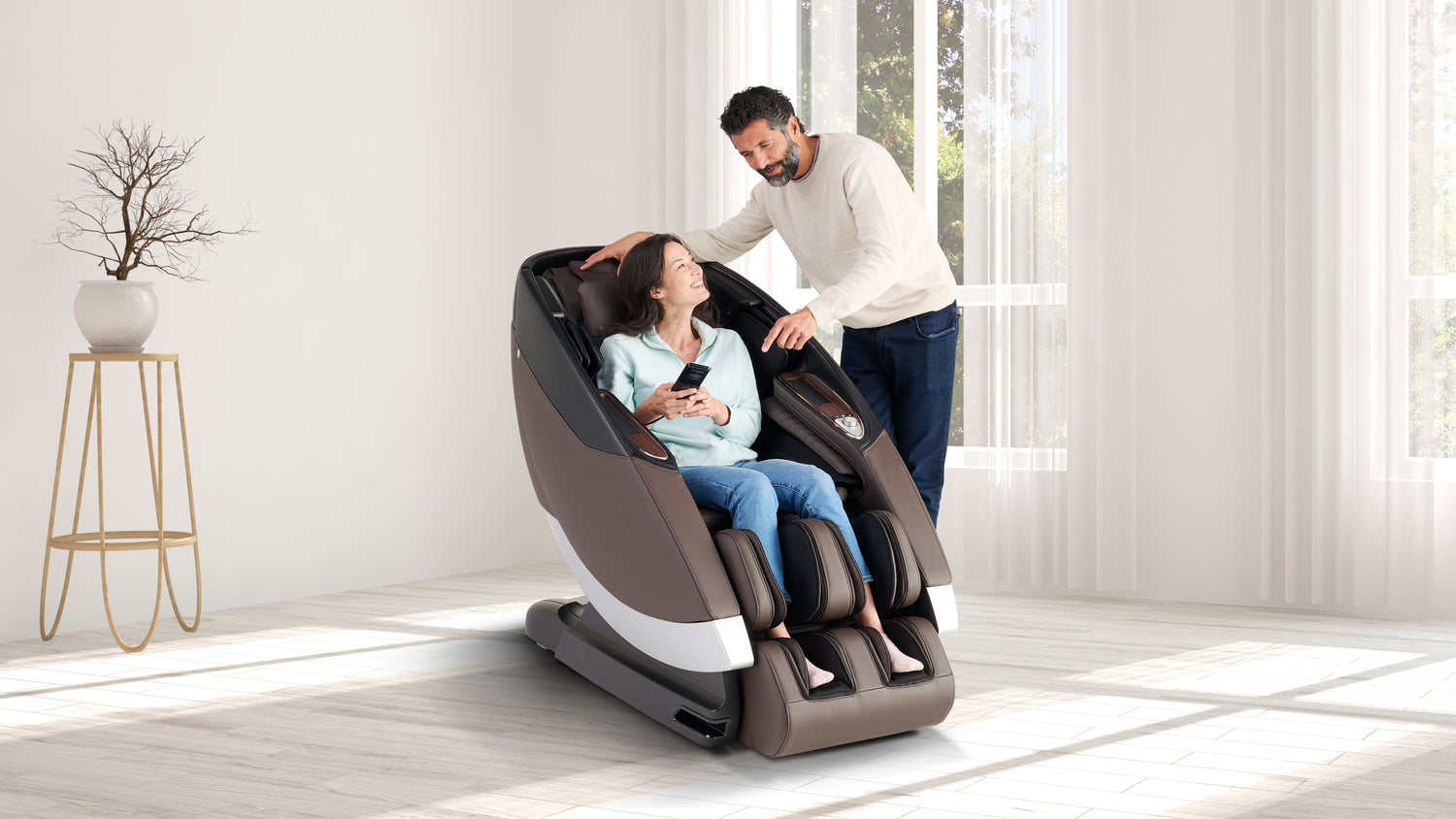 Woman sitting in a brown full-body massage chair holding remote while man stands beside her in a bright living room