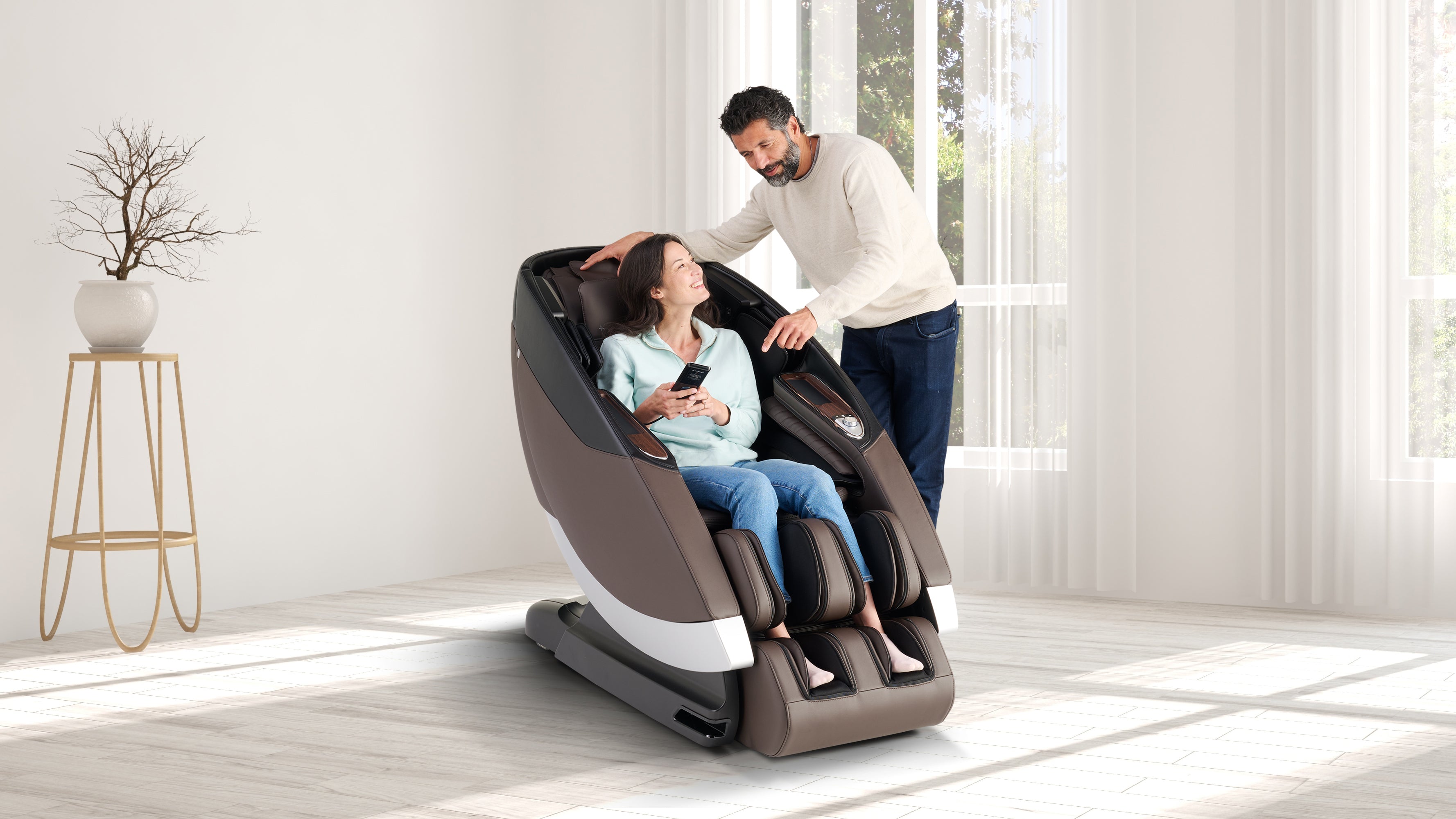Woman sitting in a brown full-body massage chair holding remote while man stands beside her in a bright living room