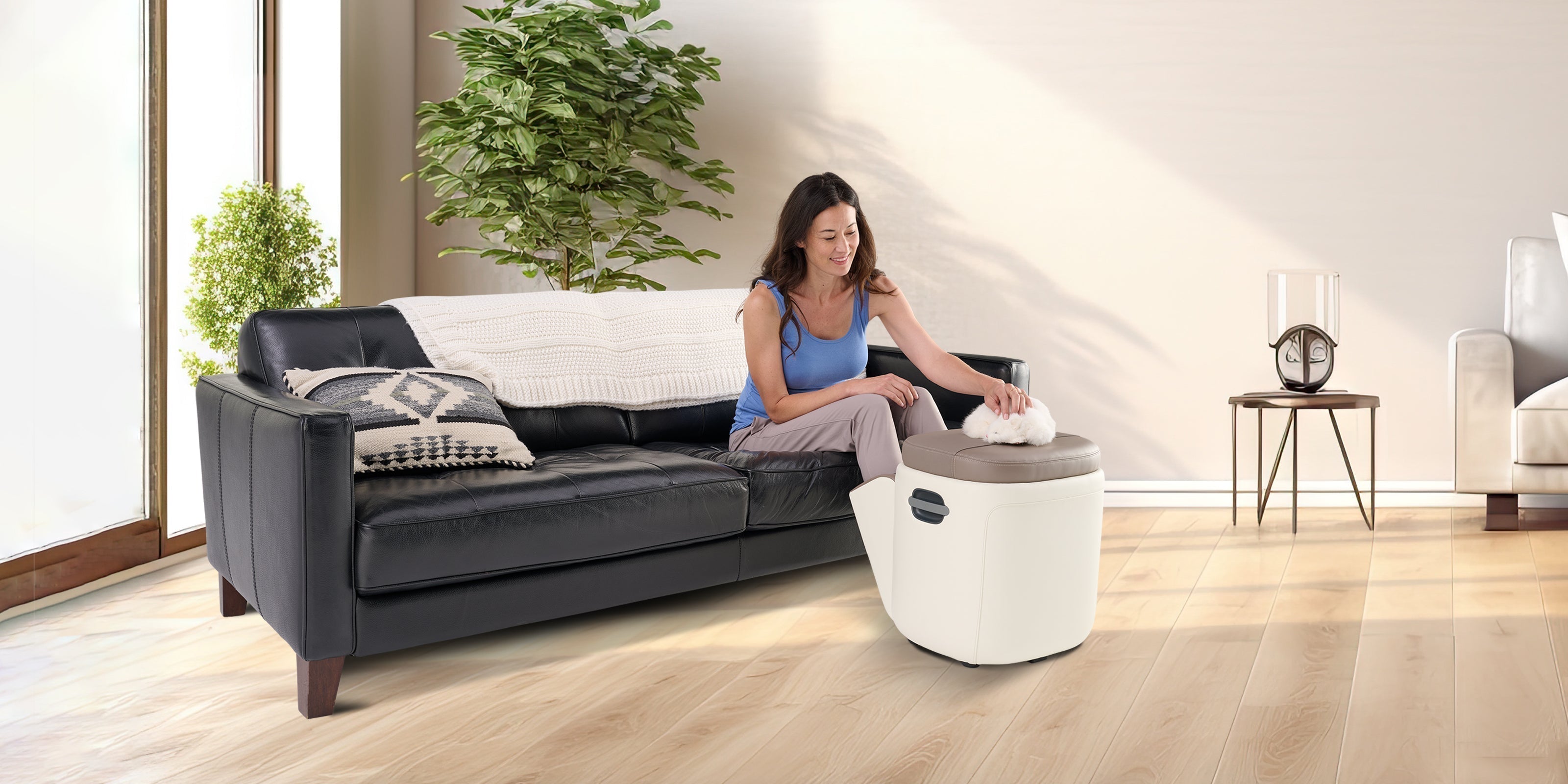 Woman sitting on a black leather sofa using a compact foot massager in a bright living room
