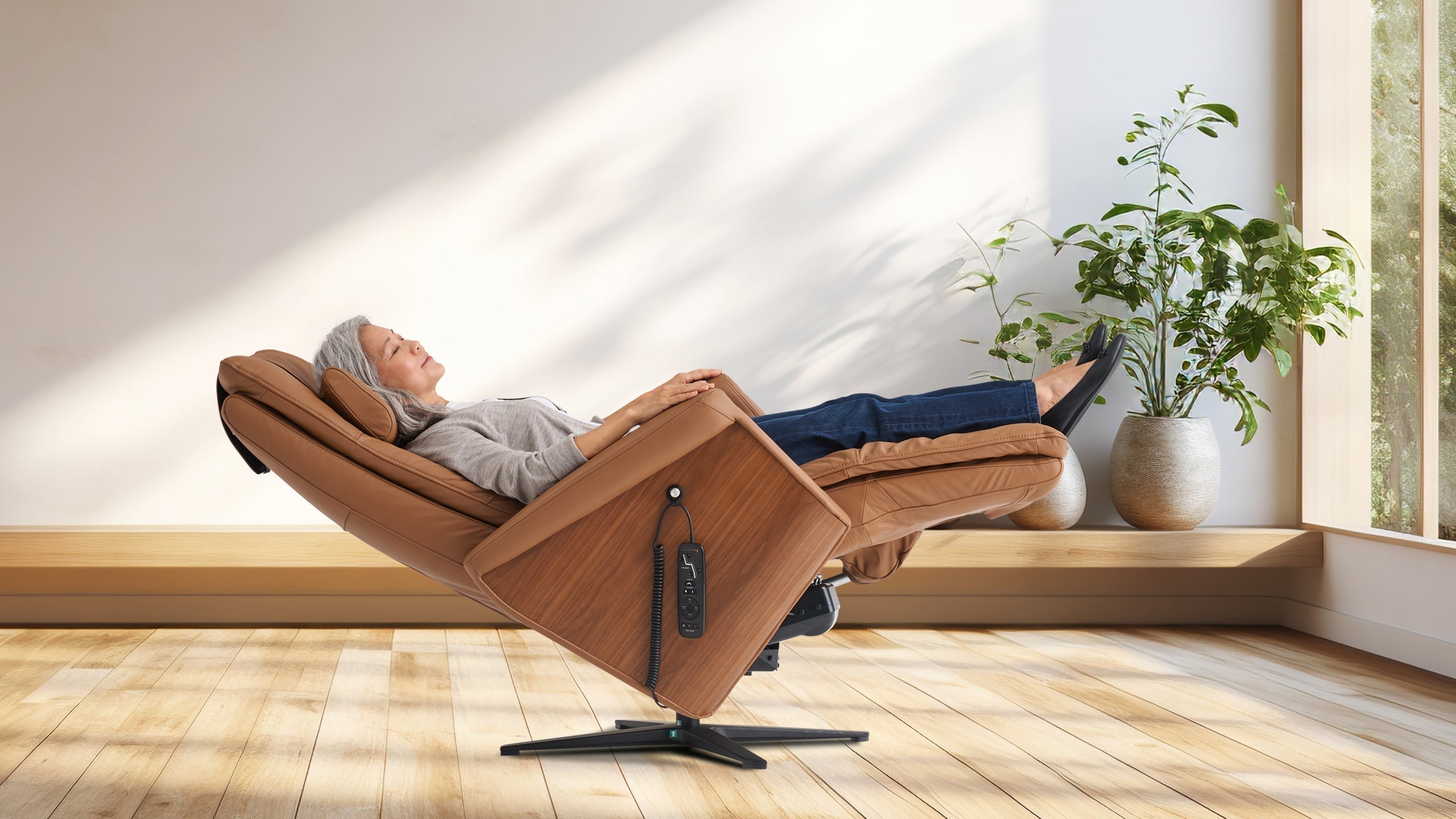 Older woman reclining in a tan leather massage recliner with legs elevated in a sunlit room with indoor plants