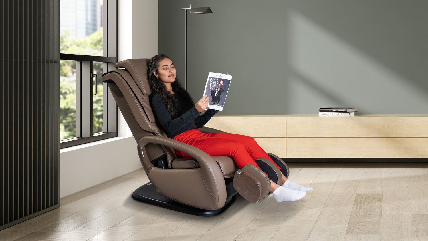 Woman reading a magazine in a brown compact massage chair next to a window in a green-walled room