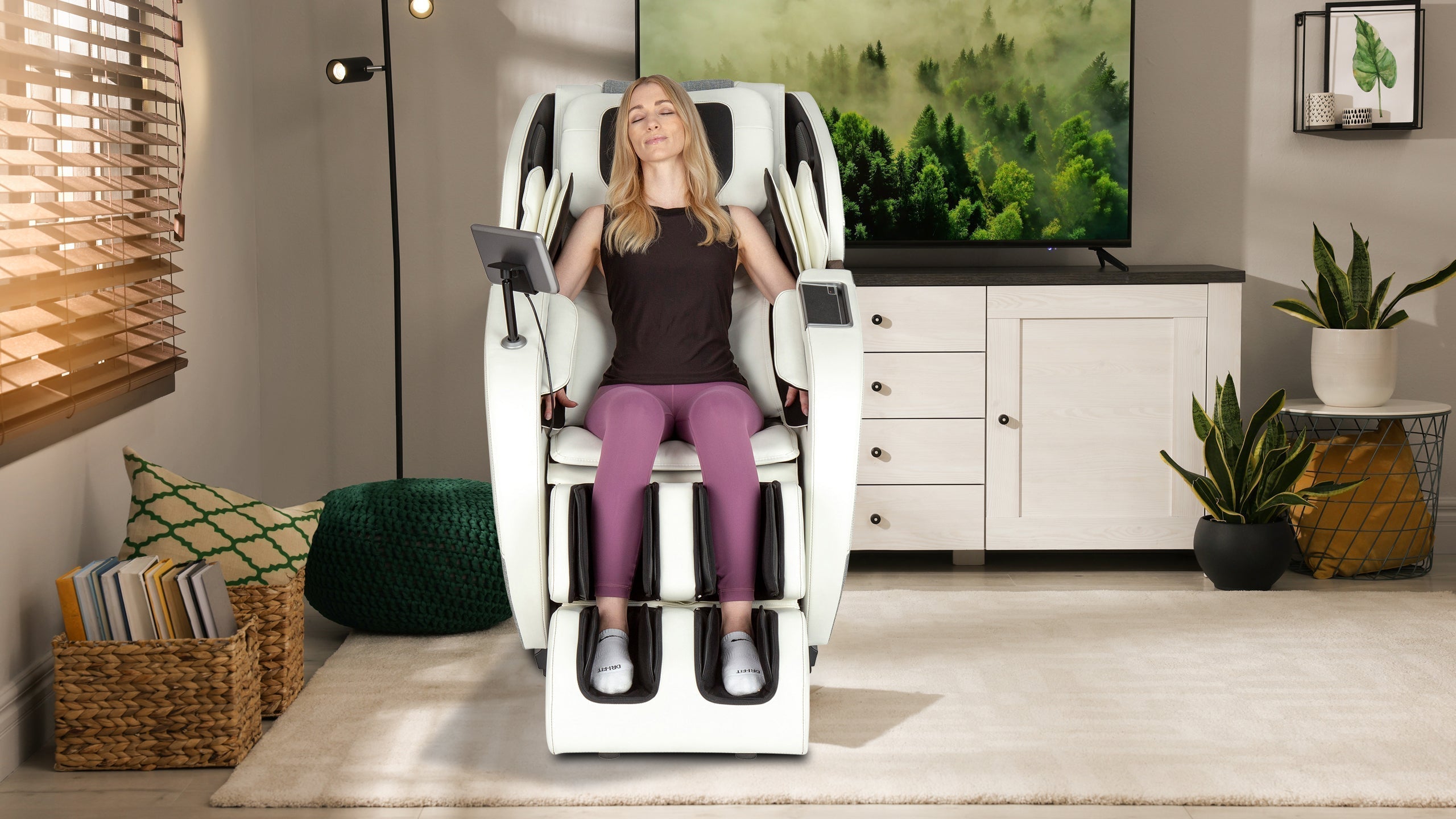 Woman relaxing with eyes closed in a white and black full-body massage chair with tablet arm in a cozy living room with TV