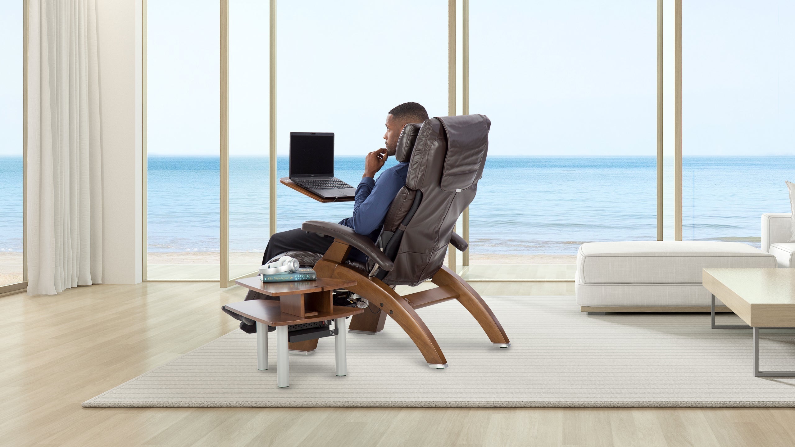 Man working on a laptop in a dark brown leather zero gravity chair with wooden side table overlooking the ocean
