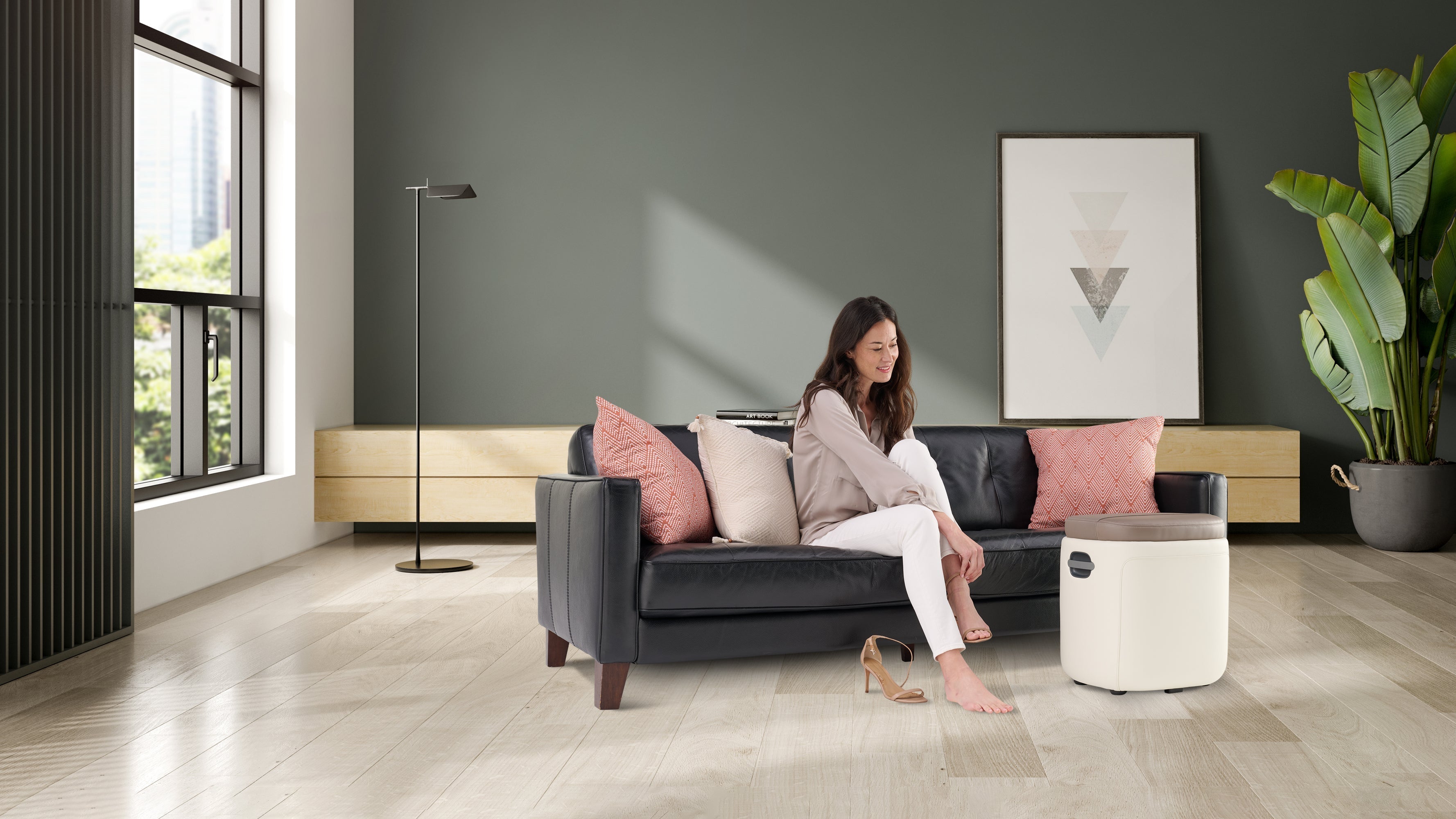 Woman sitting on black leather sofa using white compact foot massager on wooden floor in modern dark green living room