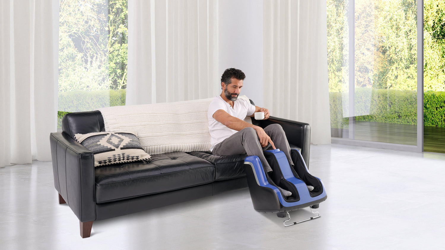 Man drinking coffee on a black leather sofa while using a blue leg and foot massager in a bright white room