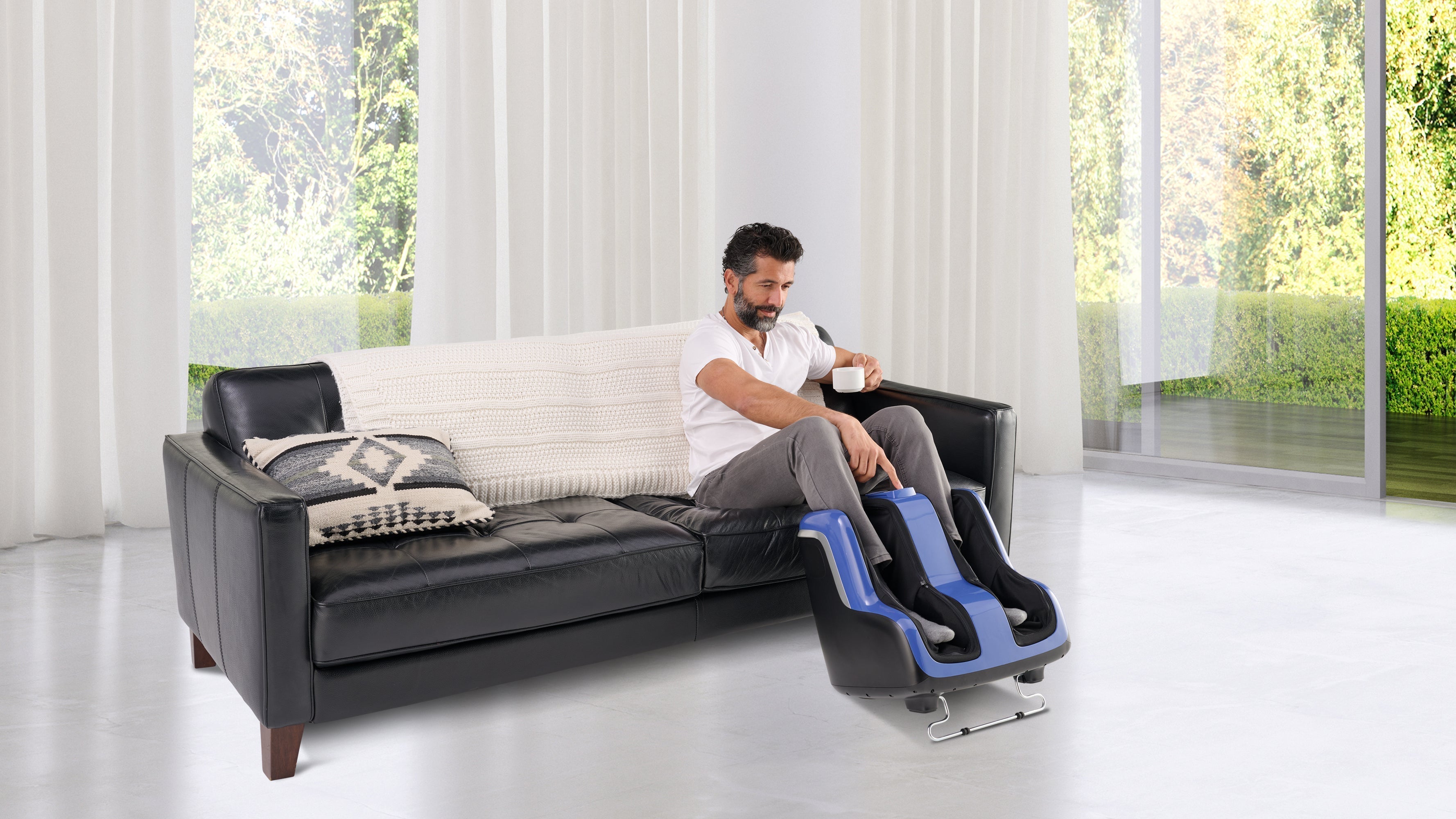 Man drinking coffee on a black leather sofa while using a blue leg and foot massager in a bright white room