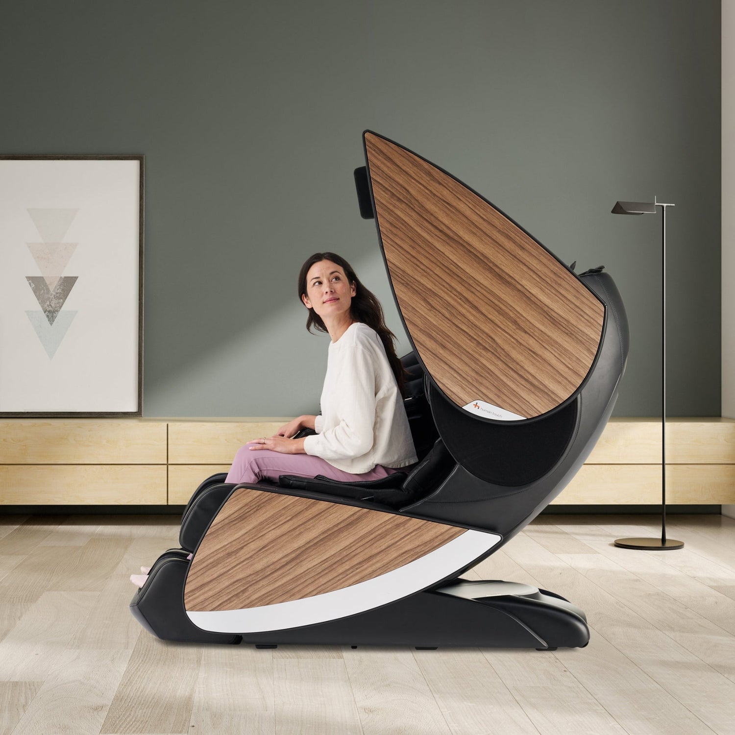 Woman sitting in a luxury black and walnut wood panel full-body massage chair with futuristic curved design in a modern room