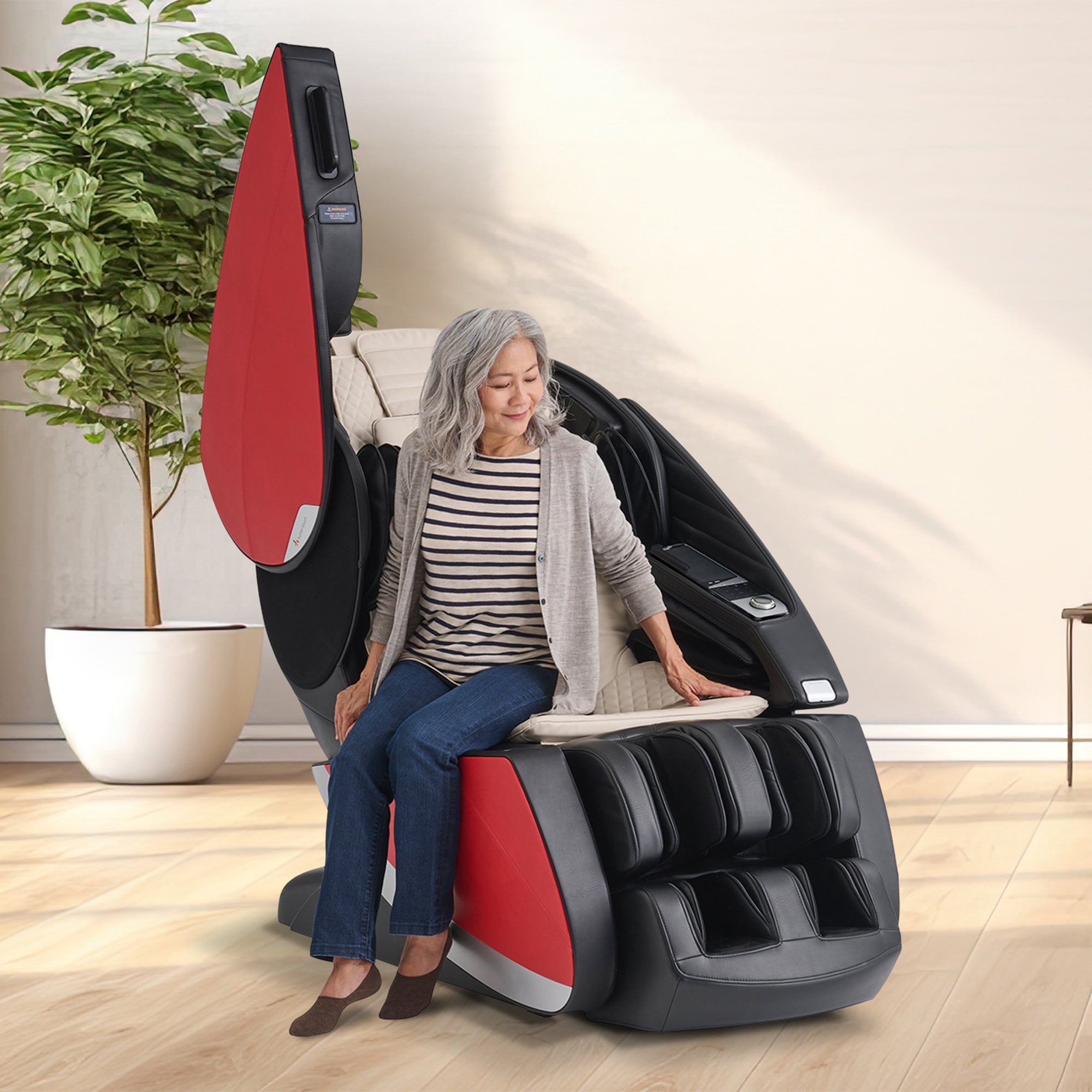 Older woman sitting in a black and red full-body massage chair with extendable tray table and open door design in a bright room