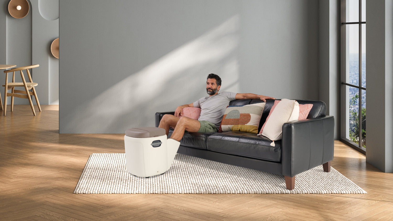 Man relaxing on a black leather sofa with a compact foot massager on the floor in a modern gray living room with city view
