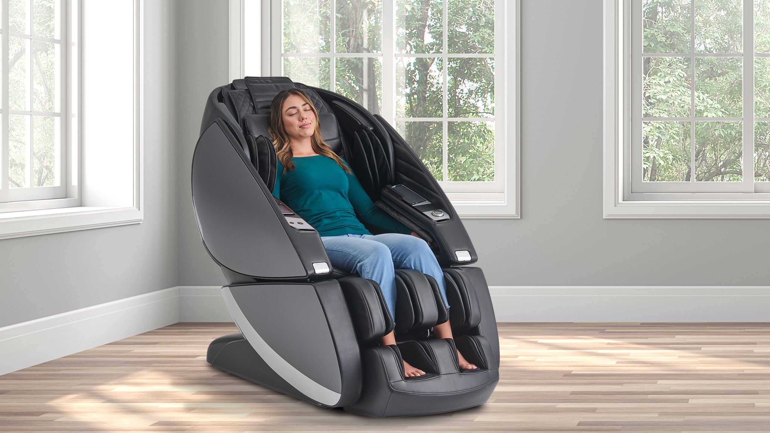 Woman relaxing with eyes closed in a black full-body massage chair in a bright room with large windows and trees outside