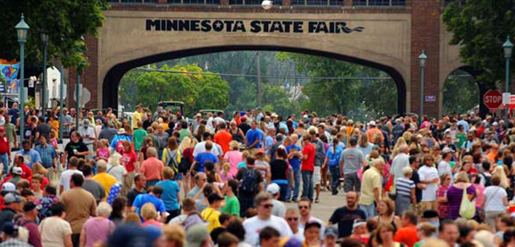 A large crowd of people walking under the brick arch of Minnesota State Fair, with the event name clearly visible above the entrance.
