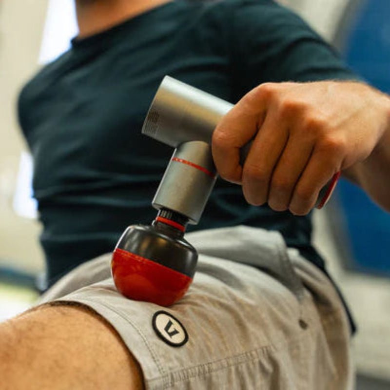 Person using a massage gun on their leg in a blurred indoor setting