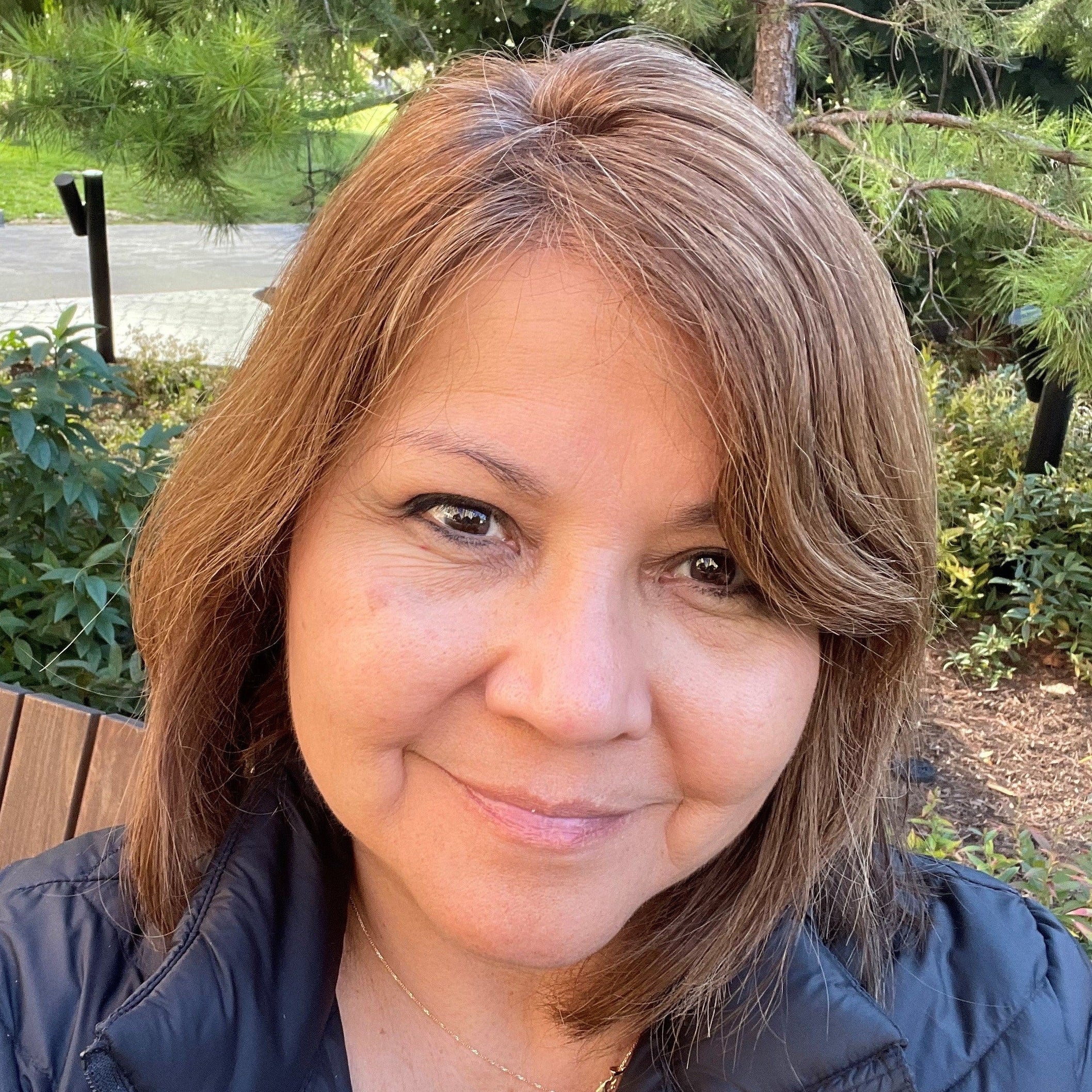 Woman with shoulder-length brown hair standing outdoors with greenery in the background