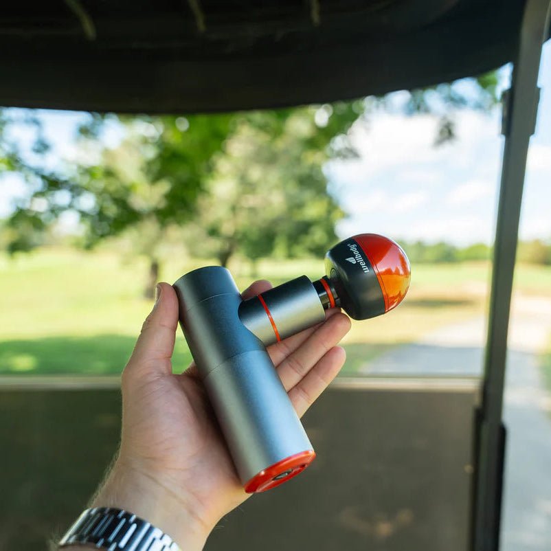 Hand holding a massage gun with a golf cart and greenery in the background