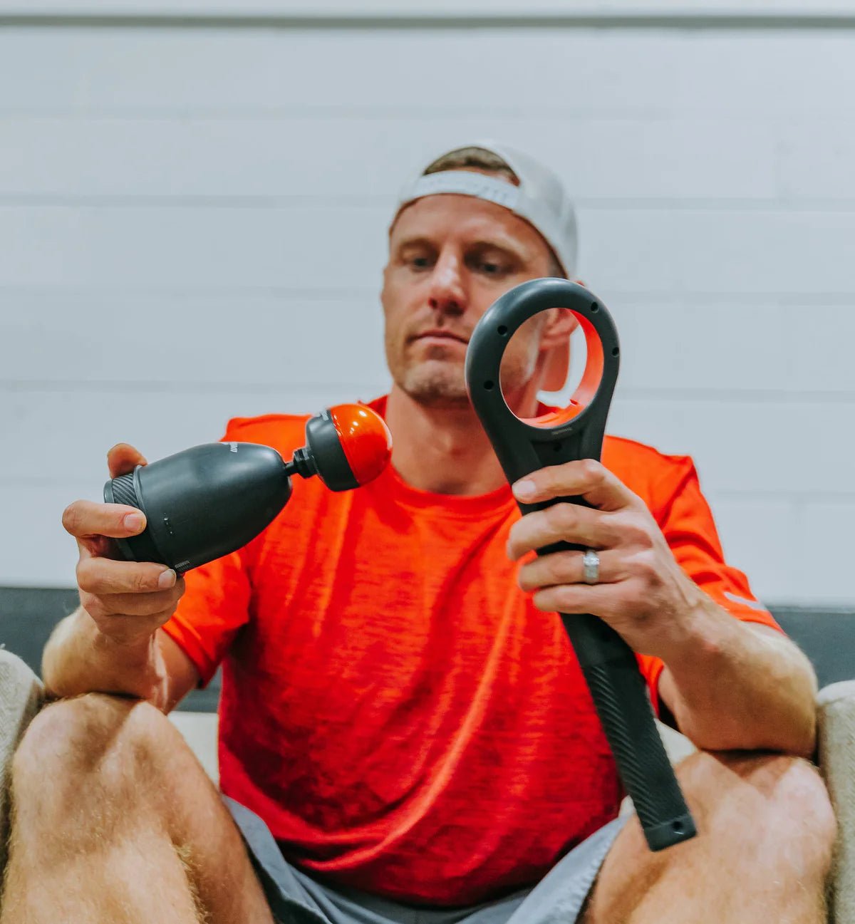 Man in orange shirt holding a massage gun and a black handheld device against a neutral background
